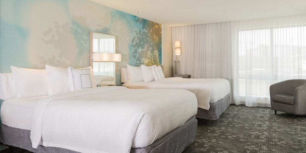 two queen beds with white bedding and light gray bed skirts in a room wiht gray patterned carpeting and chair