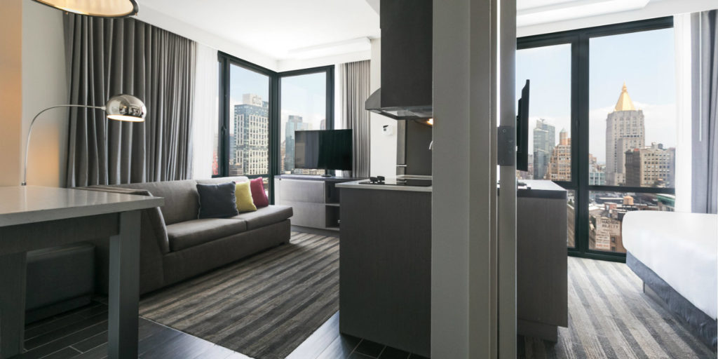 split view of a hotel room bedroom and suite area with various shades of gray furniture overlooking a large city