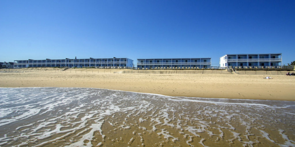 several two story blue hotels from the view of the ocean with sand, dunes and water
