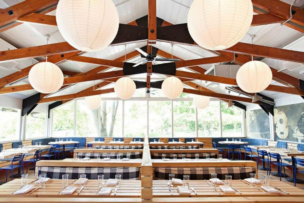 an dining area with wooden beams, round white paper lanterns and light wooden booths surrounded by large windows overlooking trees and a lawn area