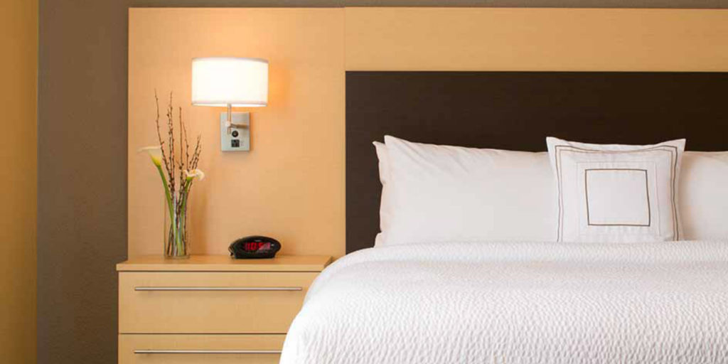 a kingsize bed with white bedding and a dark headboard next to a light wooden nightstand with alarm clock and white sconce lamp